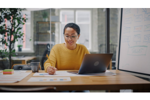Imagem de uma auxiliar administrativa trabalhando em um escritório, ela usa amarelo e a sua frente há um notebook aberto