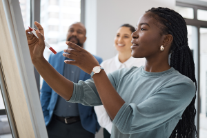 A imagem mostra uma jovem escrevendo em uma lousa, enquanto duas outras pessoas observam, representando diferentes tipos de perfil profissional. A jovem pode ser vista como a líder ativa, enquanto os outros atuam como colaboradores ou facilitadores, evide
