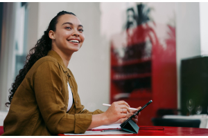 jovem sorridente sentada em uma mesa vermelha, ao ar livre, usando um tablet com caneta digital. Representando a produtividade com IA em estudos e no trabalho.