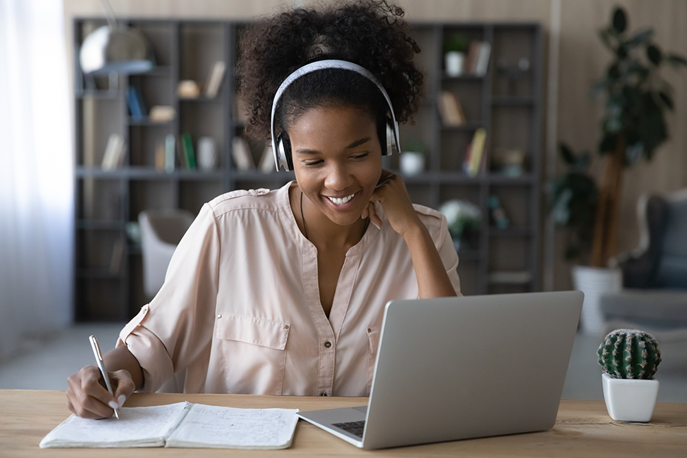 Jovem estudando com fones de ouvido, sorrindo enquanto faz anotações em um caderno e assiste a uma aula online em seu notebook — uma cena que representa perfeitamente o acesso a curso gratuito com certificado