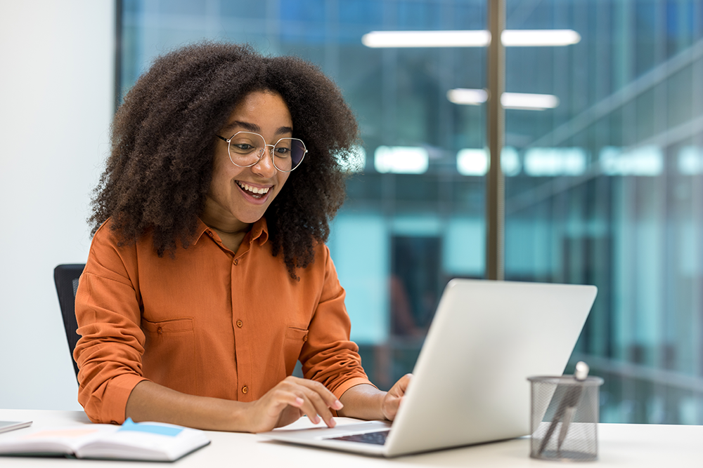 Jovem estudando Administração em frente à um notebook e sorrindo empolgada, conectada ao mercado e pronta para atuar em áreas como finanças e marketing.